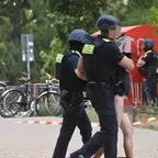 Zwei Polizisten in Schutzhelmen führen eine Person ohne Oberbekleidung an einem abgesperrten Bereich entlang, im Hintergrund Fahrräder, Bäume und ein weiterer Polizist, verschanzt hinter einem Kleidercontainer..