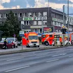 Zwei Einsatzfahrzeuge der Feuerwehr und mehrere Einsatzkräfte neben einem verunfallten roten Auto und weiter hinten, an einem weißen Auto, an einer Kreuzung vor einem Bürogebäude.