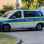 Weiß-blauer Polizeiwagen mit gelben Streifen und Blaulicht, geparkt auf einer Straße neben einem Gehweg mit Gras und Bäumen im Hintergrund.