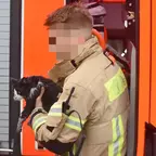 Feuerwehrmann in Schutzkleidung hält eine schwarze Katze vor einem orangefarbenen Feuerwehrfahrzeug.