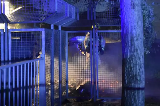 Feuerwehrleute in Schutzkleidung löschen ein Feuer hinter einem Gitterzaun bei Nacht, bläuliches Licht beleuchtet die Szene.