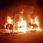 Brennendes Fahrzeug auf einer Straße bei Nacht, umgeben von Flammen und dunklem Hintergrund mit Büschen.