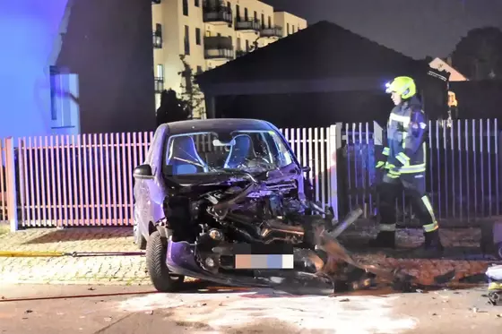 Kleiner Pkw mit stark beschädigter Front steht vor einem weißen Zaun, Feuerwehrmann in Schutzkleidung geht daneben vorbei.