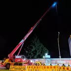 Roter Kran mit ausgefahrenem Arm, an dem eine Laterne hängt, auf einer Baustelle mit Absperrungen bei Nacht.