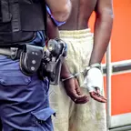 Von hinten sichtbarer Mann mit nacktem Oberkörper und Handschellen, neben ihm ein Polizist in Uniform mit Ausrüstung am Gürtel.