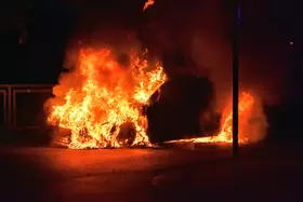 Brennendes Auto in voller Flammen, umgeben von dunkler Nacht und Rauchentwicklung.