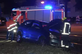 Dunkelgraues Auto mit geöffneter Motorhaube auf Grasfläche und teils im Gleisbett, Feuerwehrfahrzeug mit Blaulicht im Hintergrund, ein Feuerwehrleute in Schutzkleidung bei Nacht. Hinterm Auto zwei Mitarbeiter der BVG.