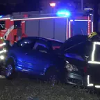 Dunkelgraues Auto mit geöffneter Motorhaube auf Grasfläche und teils im Gleisbett, Feuerwehrfahrzeug mit Blaulicht im Hintergrund, ein Feuerwehrleute in Schutzkleidung bei Nacht. Hinterm Auto zwei Mitarbeiter der BVG.