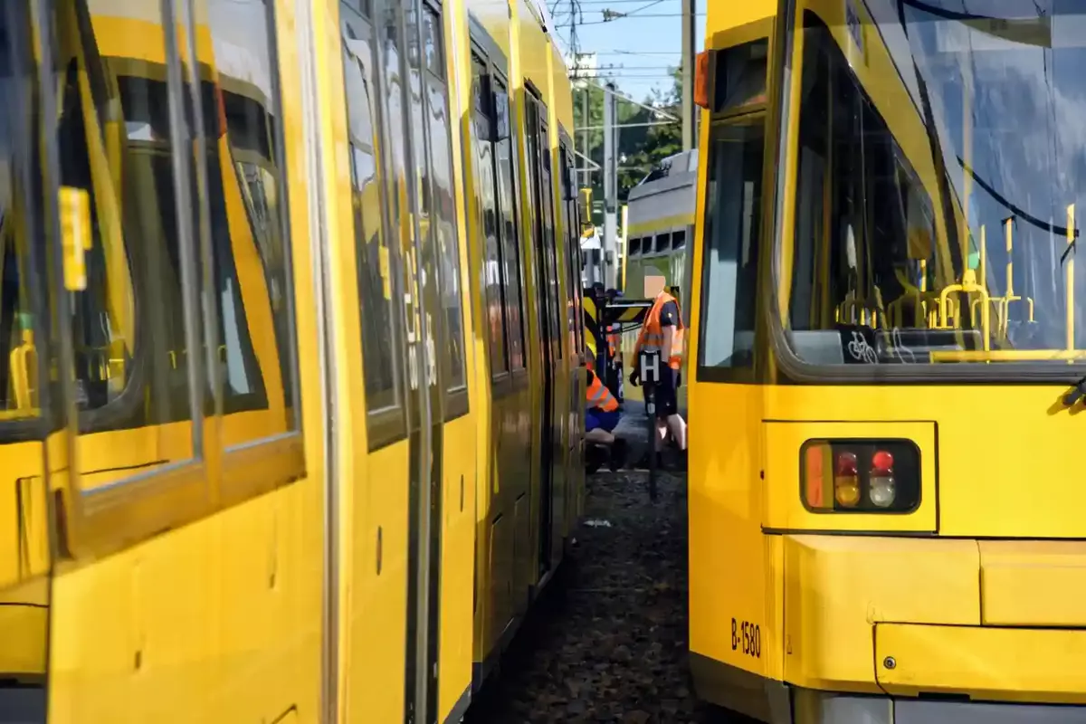 Zwei gelbe Straßenbahnen stehen auf Gleisen, zwischen ihnen sind zwei Personen in orangefarbenen Warnwesten zu sehen.