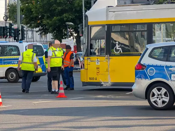 Mehrere Personen in Warnwesten stehen neben einer gelb-weißen Straßenbahn mit sichtbaren Beschädigungen an der Seite. Rechts und links sind Polizeifahrzeuge in blau-weiß zu sehen. Im Vordergrund stehen zwei orangefarbene Verkehrskegel.