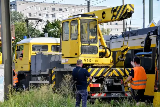 Gelber Kranwagen mit schwarzem Warnmuster, zwei Männer in Warnwesten stehen daneben, im Hintergrund Wohngebäude und Oberleitungen.