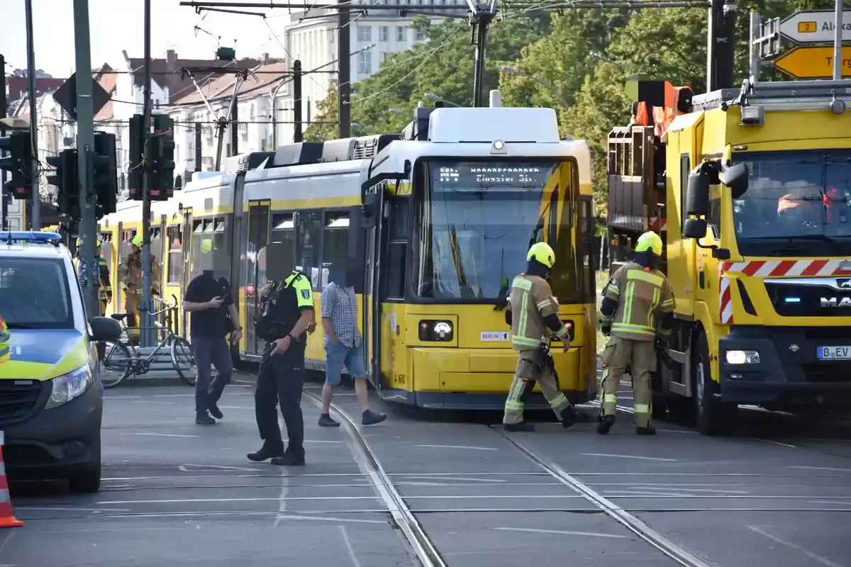 Eine entgleiste gelbe Straßenbahnen steht an Gleisen in einer Stadtstraße, Feuerwehrleute in Schutzkleidung und Polizisten sind vor Ort, Passanten gehen vorbei.