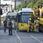 Eine entgleiste gelbe Straßenbahnen steht an Gleisen in einer Stadtstraße, Feuerwehrleute in Schutzkleidung und Polizisten sind vor Ort, Passanten gehen vorbei.