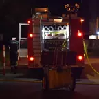 Rückansicht eines roten Feuerwehrautos bei Nacht mit geöffnetem Geräteraum und gelbem Schlauch, daneben eine Person in dunkler Kleidung.