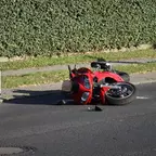 Rotes Motorrad liegt auf der Seite auf einer Asphaltstraße neben einem Gehweg mit Hecke im Hintergrund.