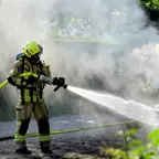 Feuerwehrmann in gelber Schutzkleidung und Helm spritzt Wasser aus Schlauch in rauchiger Umgebung.