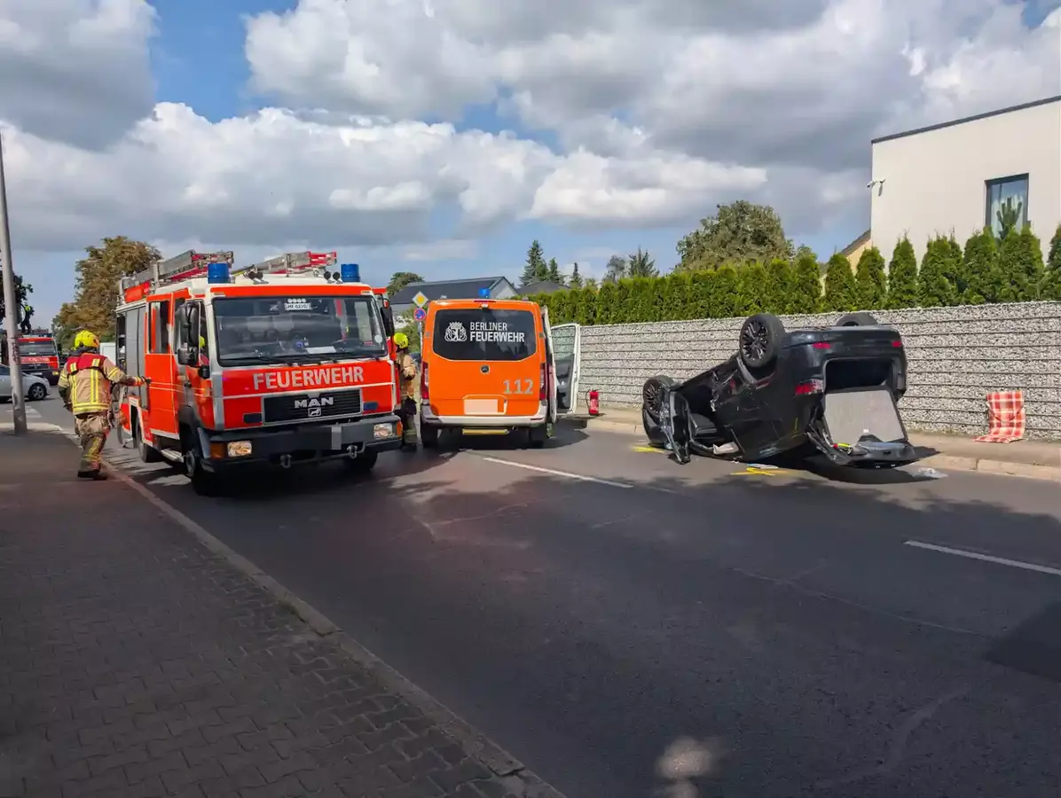 Ein schwarzes Auto liegt auf dem Dach auf einer Straße, daneben stehen ein rotes Feuerwehrfahrzeug und ein Notarzteinsatzfahrzeug der Berliner Feuerwehr, Feuerwehrleute sind vor Ort.