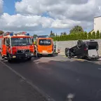 Ein schwarzes Auto liegt auf dem Dach auf einer Straße, daneben stehen ein rotes Feuerwehrfahrzeug und ein Notarzteinsatzfahrzeug der Berliner Feuerwehr, Feuerwehrleute sind vor Ort.