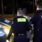 Drei Polizisten in dunkler Uniform führen eine Person mit hochgekrempelten Ärmeln zum Einsatzfahrzeug bei Nacht.