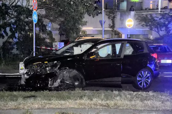 Schwarzes Auto mit stark beschädigter Front und geöffneter Fahrertür steht nachts auf einer Straße vor einem Wohnhaus.