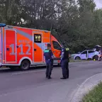 Zwei Polizisten stehen auf einer Straße neben einem orange-roten Rettungswagen mit der Aufschrift 'NOTRUF 112', im Hintergrund ein weißes Auto abseits der Fahrbahn und ein Feuerwehrfahrzeug.