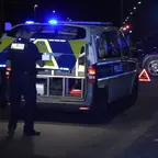 Polizist steht neben einem geöffneten Polizeifahrzeug mit eingeschaltetem Blaulicht auf einer Straße bei Nacht, im Hintergrund ein weiteres Fahrzeug mit Warnblinker und Warndreieck.