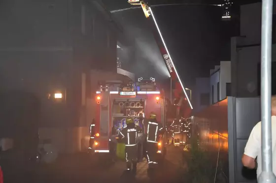 Feuerwehrleute in Schutzkleidung vor einem Feuerwehrfahrzeug, dahinter ein Weiteres mit ausgefahrener Leiter in einer dunklen Gasse bei Nacht.