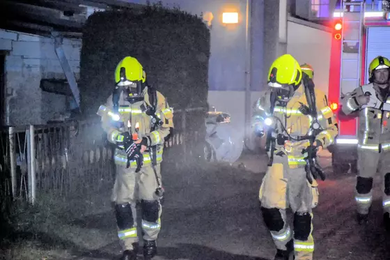 Zwei Feuerwehrleute in gelben Helmen und Schutzanzügen gehen nachts auf einer Straße, eine dritte Person mit reflektierender Kleidung steht daneben.