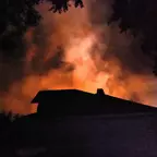 Dunkle Silhouette eines Hauses vor orangefarbenem Rauch oder Feuer, umgeben von Bäumen in der Nacht.