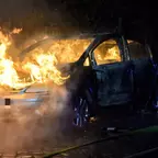 Brennendes Auto mit Flammen aus Motorraum und Innenraum, umgeben von Rauch in der Nacht.