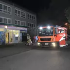 Rotes Feuerwehrfahrzeug mit eingeschalteten Scheinwerfern steht nachts vor einem Gebäude mit bunter Wandmalerei, Feuerwehrleute in Schutzkleidung sind zu sehen.