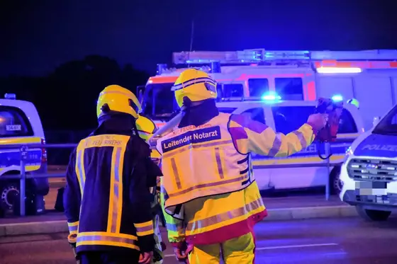 Zwei Einsatzkräfte in gelben Helmen und Warnwesten, eine mit der Aufschrift 'Leitender Notarzt', stehen vor Einsatzfahrzeugen mit Blaulicht.