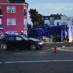 Schwarzes Auto mit beschädigter Front steht vor umgekipptem blauen Lkw auf Straße, Feuerwehrleute und Fahrradfahrer im Hintergrund.