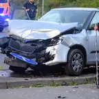 Silberner Pkw mit stark beschädigter Front steht auf Mittelinsel und Verkehrsschild, zwei Polizisten und ein Rettungswagen im Hintergrund.