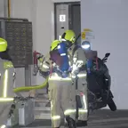 Drei Feuerwehrleute in gelben Helmen und reflektierender Schutzkleidung stehen vor einem Gebäude mit offener Tür und Briefkästen, ein Moped steht daneben.