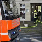 Feuerwehrauto der Berliner Feuerwehr mit eingeschalteten Blaulichtern vor einem Gebäude, Feuerwehrleute mit Schutzkleidung und gelben Warnstreifen führen Schläuche ins Gebäude.