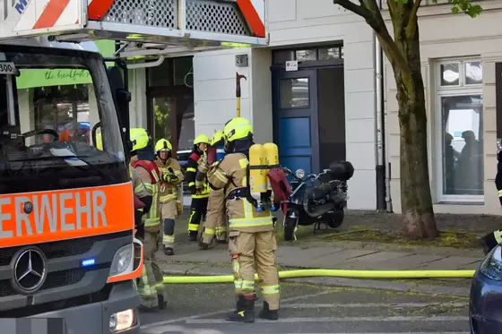 Mehrere Feuerwehrleute in gelber Schutzkleidung und Helmen stehen vor einem Gebäude, eine Drehleiter ist links im Bild zu sehen.