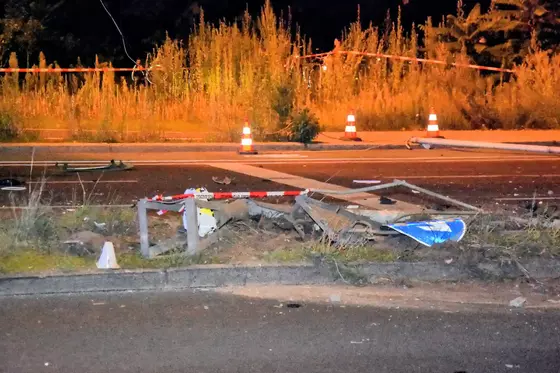 Zerstörtes Verkehrsschild und ein Ampelmast liegt auf dem Boden an einer Straße, im Hintergrund mehrere orange-weiße Leitkegel und hohe Vegetation.