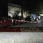 Absperrband mit der Aufschrift 'FEUERWEHRSPERRZONE' vor geparkten Autos und zwei Polizisten auf einem gepflasterten Weg bei Nacht.