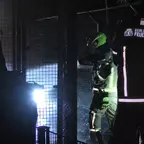 Zwei Feuerwehrleute in Schutzkleidung und Helmen stehen vor einem Gitter, Lichtstrahl beleuchtet die Szene von links.