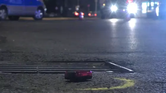 Rote Fahrrad-Rücklicht liegt auf der Straße vor einem Gullydeckel, im Hintergrund unscharfe Autoscheinwerfer und Fahrzeuge.