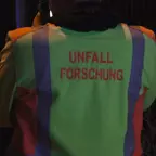 Rückansicht einer Person in gelb-grüner Warnweste mit roten Streifen und der Aufschrift 'UNFALL FORSCHUNG'.