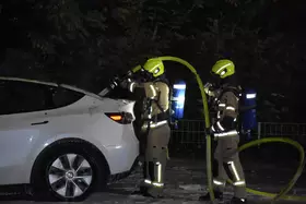 Zwei Feuerwehrleute in gelben Helmen und Schutzanzügen stehen neben einem weißen Auto und halten einen gelben Schlauch.