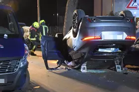Auf dem Dach liegender silberner Pkw mit geöffneter Fahrertür, daneben zwei Feuerwehrleute in gelben Helmen und Schutzkleidung auf einer Straße bei Nacht.