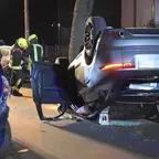 Auf dem Dach liegender silberner Pkw mit geöffneter Fahrertür, daneben zwei Feuerwehrleute in gelben Helmen und Schutzkleidung auf einer Straße bei Nacht.