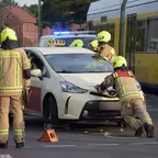 Mehrere Feuerwehrleute in gelben Helmen und Schutzkleidung schieben ein Taxi auf einer Straße neben einer gelben Straßenbahn.