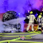 Feuerwehrleute löschen nachts einen brennenden Pkw mit geöffnetem Motorraum, umgeben von Rauch und mehreren Schläuchen auf der Straße.