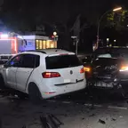 Zwei beschädigte Autos stehen nachts auf der Straße, ein weißer VW und ein schwarzes Fahrzeug mit eingeschaltetem Scheinwerfer, im Hintergrund ein Einsatzfahrzeug mit Blaulicht.