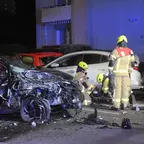 Zerstörter schwarzer Pkw mit eingedrückter Front, drei Feuerwehrleute in gelben Helmen und Schutzkleidung arbeiten an der Unfallstelle bei Nacht.