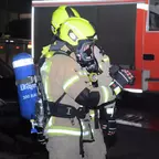 Zwei Feuerwehrleute in gelben Helmen und Atemschutzgeräten, einer hält eine Wärmebildkamera, im Hintergrund ein rotes Feuerwehrfahrzeug.